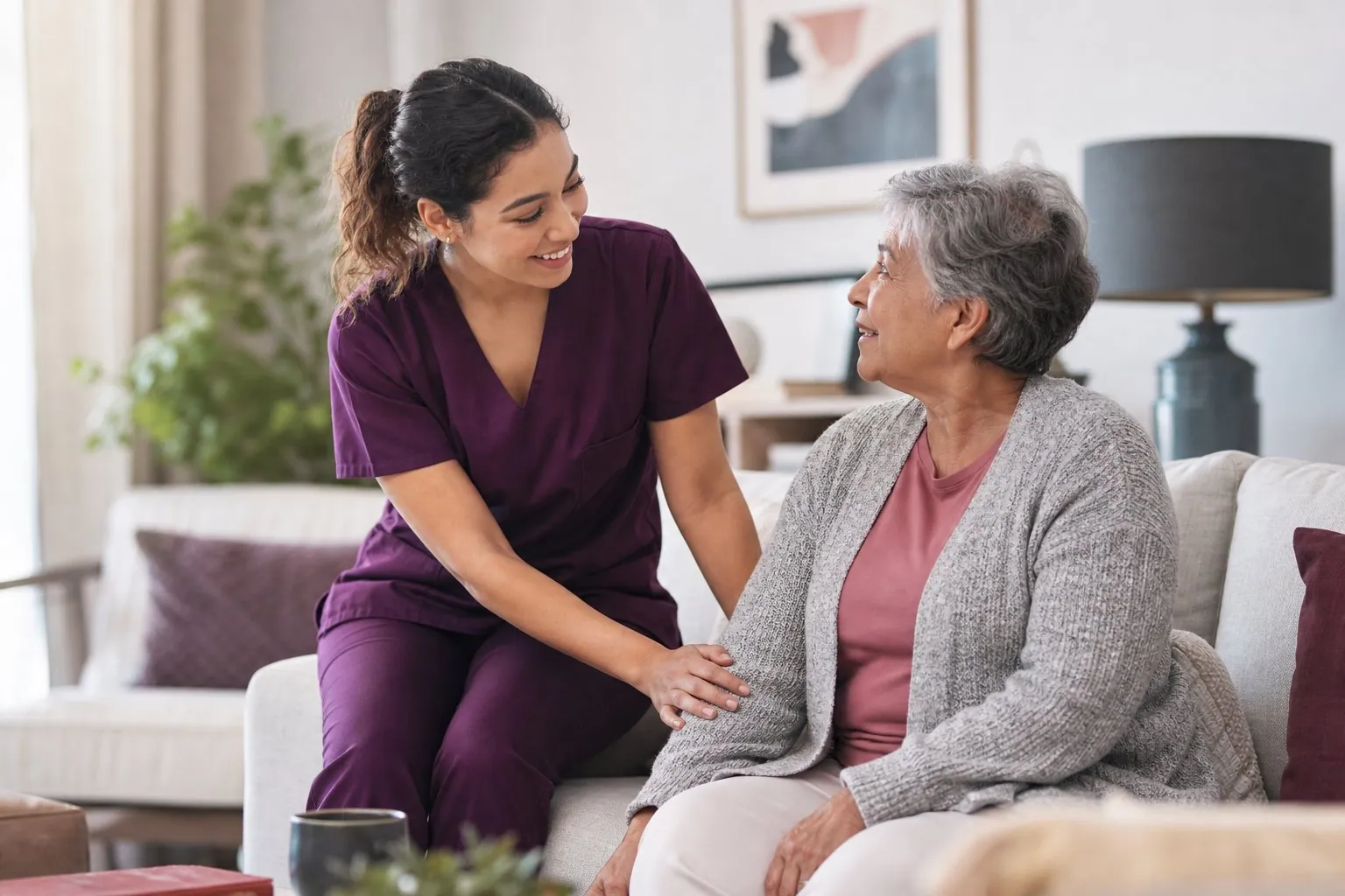 A home health aide in scrubs warmly assisting a senior woman in her home